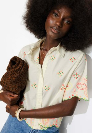 Femme aux cheveux afro naturels portant une chemise crème brodée et un jean bleu, tenant un tissu marron à motifs, debout devant un fond blanc.
