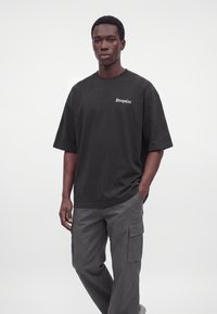 Jeune homme portant un t-shirt noir oversize avec le texte "Dropsize" et un pantalon cargo gris, debout devant un fond blanc uni.