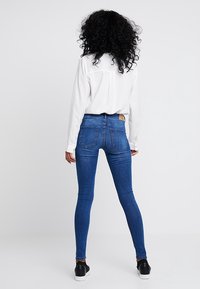 Jeans ajustados de color azul con una cintura de altura media, que presentan un diseño clásico de cinco bolsillos, detalle de costura y una etiqueta de marca sutil en la parte trasera.