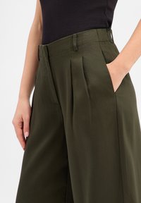 Olive green high-waisted trousers with a smooth texture, featuring front pleats and side pockets. The fabric appears lightweight and tailored.