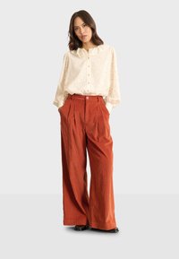 Blusa in tessuto cremoso con colletto arricciato e chiusura a bottoni, abbinata a pantaloni in corduroy ruggine a gamba larga con tasche laterali e pieghe.