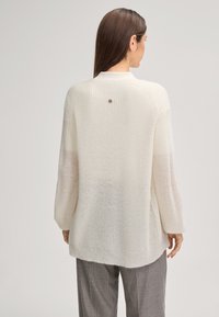 Donna che indossa un maglione lavorato a maglia bianco e largo con un piccolo emblema rotondo sulla parte superiore della schiena, abbinato a pantaloni a quadri grigi, girata di spalle.