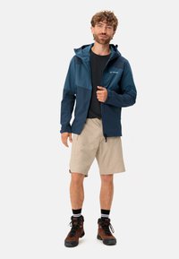 Veste imperméable bleu et bleu foncé avec capuche, fermeture éclair et design de panneaux contrastants, portée avec un short léger et des bottes de randonnée.