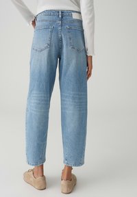 Persona che indossa jeans azzurri a gamba dritta, sneakers beige e una maglia bianca a maniche lunghe, mostrata di spalle su uno sfondo semplice.