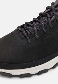 Černé semišové tenisky s texturovanými tkaničkami, polstrovaným límcem a kontrastní bílou mezipodešví s robustní černou podešví pro lepší trakci.