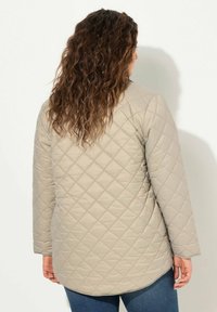 Chaqueta acolchada beige claro con un patrón de diamantes, mangas largas y un dobladillo redondeado, combinada con jeans de mezclilla azul.