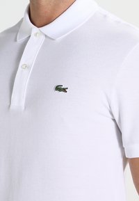 Polo blanc en coton texturé, avec un col classique, deux boutons et un petit logo crocodile vert brodé sur la poitrine.