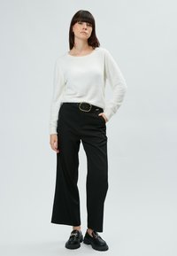 Maglione bianco a maniche lunghe, con texture, abbinato a pantaloni neri larghi, con una cintura con fibbia dorata, e mocassini neri chunky.