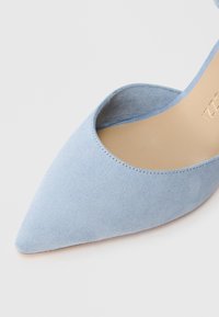 Απαλή μπλε σουέτ γόβα με αιχμηρή μύτη και κοπή λεπτομέρεια, λεία υφή και μπεζ σόλα. Διαθέτει ελάχιστη ραφή.