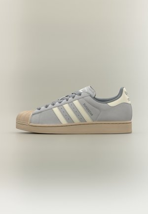 Szary but sportowy Adidas Superstar z kremowym noskiem, trzema białymi paskami, beżową podeszwą i napisem „Superstar” po boku, pokazany z widoku z lewej strony.