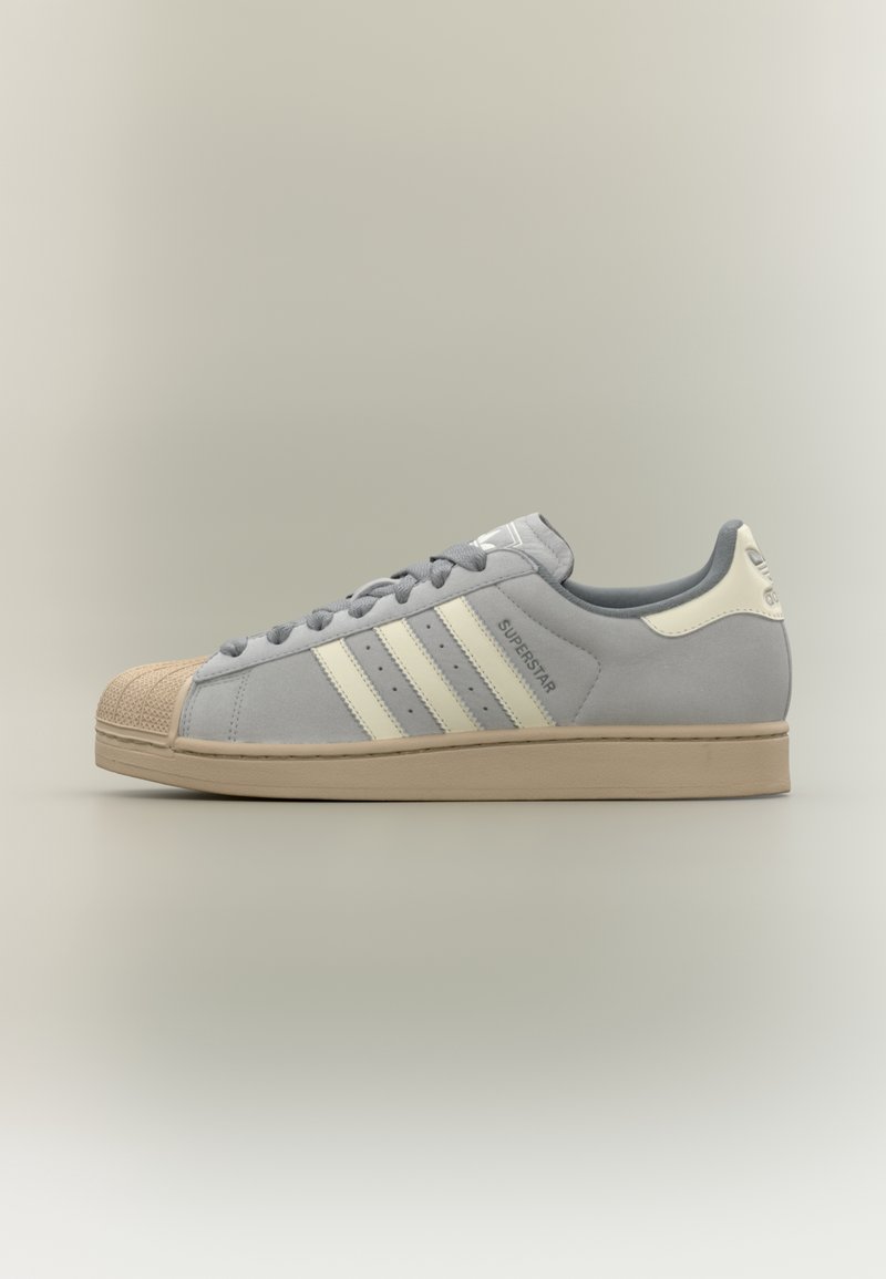 Baskets Adidas Superstar grises avec bout crème, trois bandes blanches, semelle beige et inscription « Superstar » sur le côté, vues de profil gauche.