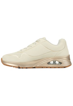 Sneaker beige chiaro con una tomaia sintetica liscia, accenti perforati, dettagli dorati e una soffice imbottitura d'aria visibile nella suola. Suola in gomma.