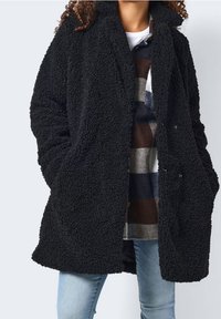 Svart, fluffig ullkappa med lös passform, knappstängning och två framfickor. Bärs över en mönstrad skjorta och ljusblå jeans.