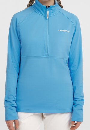 Person iført en lyseblå O'Neill langærmet pullover med kvart-zip og hvide bukser på en ensfarvet baggrund.