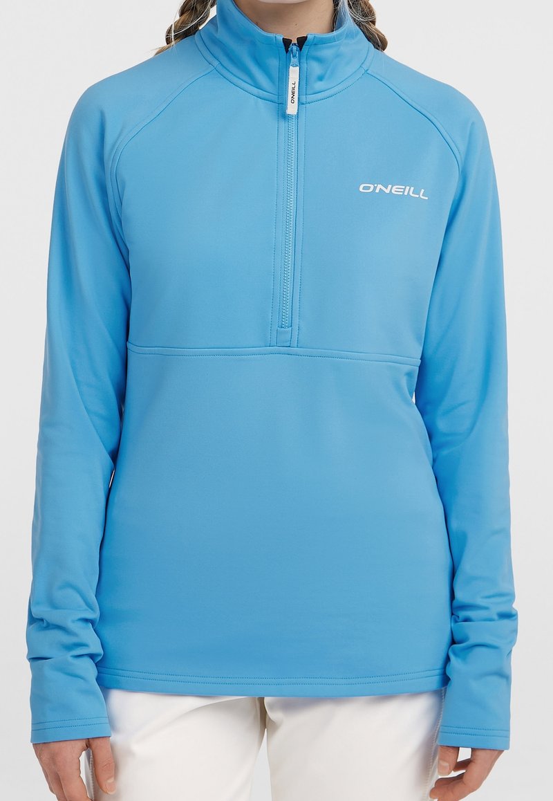 Personne portant un pull à manches longues O'Neill bleu clair avec une fermeture éclair quart de tour et un pantalon blanc sur un fond uni.
