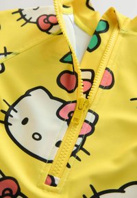 Sárga fürdőruha, amely Hello Kitty mintával rendelkezik, fekete körvonalakkal és rózsaszín díszítésekkel, valamint funkcionális elülső cipzárral. Simább, rugalmas anyag.