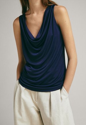 Vrouw draagt een mouwloze donkerblauwe top met gedrapeerde hals en lichtbeige plissébroek met hoge taille, handen in de zakken.