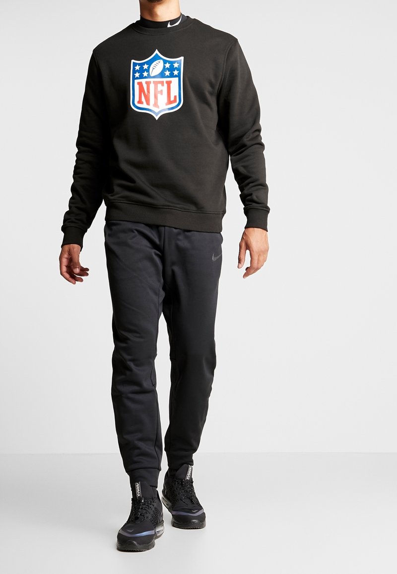 Felpa nera con logo NFL colorato, abbinata a pantaloni sportivi neri e sneakers nere con dettagli testurizzati.