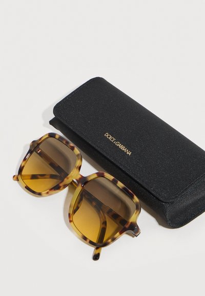 Γυαλιά ηλίου με τετράγωνο σκελετό σε χελώνα και καφέ φακούς με διαβάθμιση, δίπλα σε μαύρη ορθογώνια θήκη Dolce & Gabbana με χρυσά γράμματα.