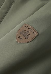 Olivno zelena tkanina z gladko teksturo, ki ima rjavo usnjeno nalepko z odtisnjenimi drevesi in besedilom "N° 18.25 W NMT."
