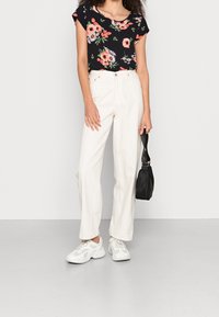 Top negro floral con mangas cortas, pantalones blancos de cintura alta y zapatillas blancas. Sosteniendo un pequeño bolso negro, de pie sobre un fondo claro.