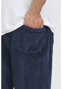 Kordurové kalhoty tmavě modré barvy s vertikálními žebernými pruhy, s našitou kapsou. Ruka spočívá v kapse. Nad nimi je vidět bílé tričko.