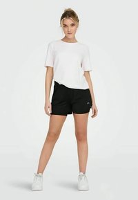 Noor noor seisab käed lõdvestatult, kandes valget T-särki, musti lühikesi pükse ja valgeid spordijalanõusid, lihtsa helepruuni tausta ees.