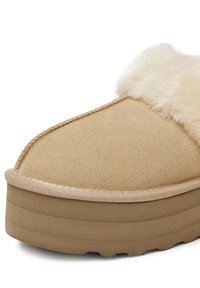 Béžové semišové pantofle s tlustou, strukturovanou podešví, s huňatým krémovým kožíškem podél horního okraje a viditelnými švy.