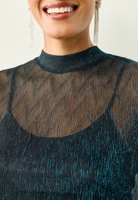 Prozirna crna bluza sa metalnim nitima u boji potočnice, visokim vratom i teksturiranim uzorkom. Ima slojeviti dizajn i elegantne duge naušnice.