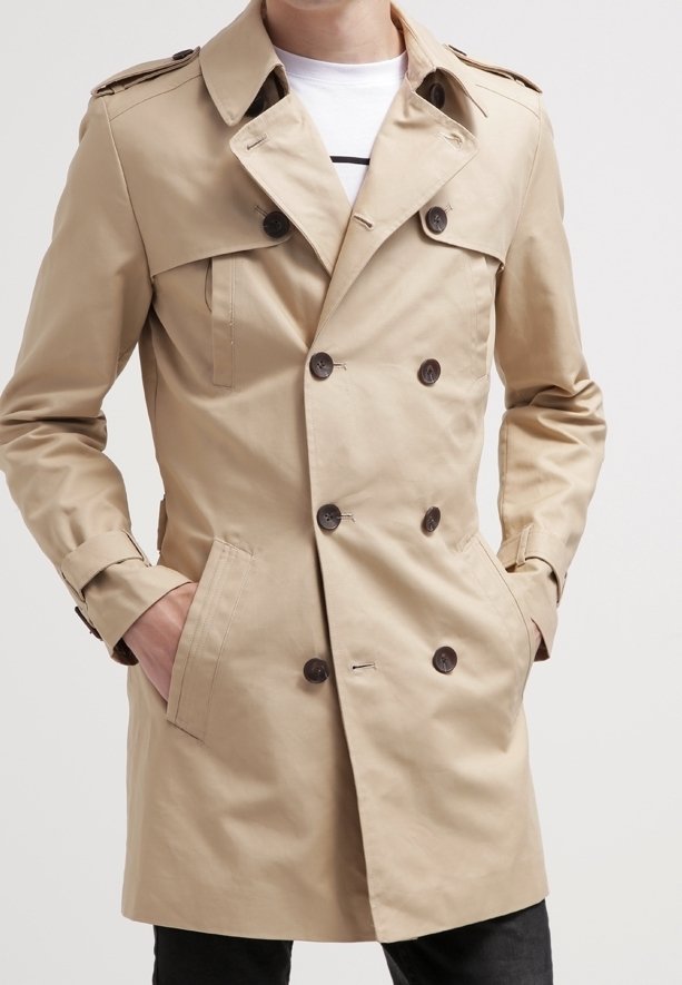 Trench-coat beige à double boutonnage avec des boutons foncés, des épaulettes et des poches latérales, porté sur une chemise blanche, mains dans les poches.