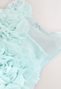 Robe en tissu léger bleu clair avec détails de volants superposés et fines bretelles sur un fond uni clair.