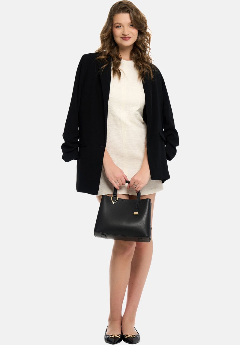 Blazer nero su un vestito crema, abbinato a una borsa nera e a scarpe flats nere con accenti dorati. Il tessuto strutturato e la vestibilità su misura sono notevoli.