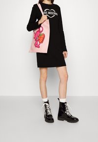 Kvinna i svart långärmad klänning med texten "LOVE MOSCHINO", bärande på en rosa toteväska med en tecknad mus och jordgubbe, iförd svarta snörstövlar.