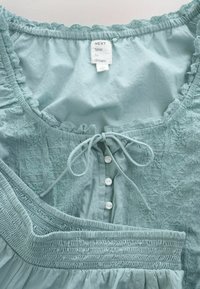 Robe sans manches bleu clair avec bordure en dentelle, boutons sur le devant, lien à l'encolure et smocks élastiques à la taille et aux poignets.