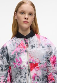 Giacca bomber con un design grafico riflettente e multicolore che presenta motivi rosa, grigi e rossi. Ha una chiusura frontale con cerniera e un colletto nero.