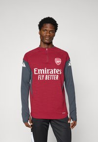 Maillot de sport bordeaux avec col à fermeture éclair sur le quart, manches grises et logo blanc "Emirates FLY BETTER", présentant des motifs texturés sur le tissu.