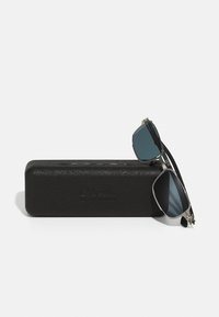 Γυαλιά ηλίου με γκρι φακούς και ασημένιο μεταλλικό σκελετό, εναποκειμένα σε μια μαύρη, υφασματισμένη σκληρή θήκη με ανάγλυφο λογότυπο.