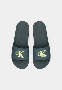 Sandales de plage bleu foncé avec une large sangle portant un logo "CK" jaune, fabriquées en matériau en caoutchouc doux pour le confort, avec un design de semelle plate.