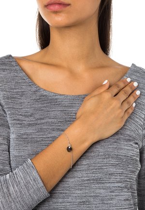 Sølvarmbånd med en sort hjerteformet charme. Har en delikat kæde og en simpel lås. Båret på et bar håndled mod en grå bluse.