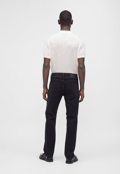 Homme portant un polo blanc à manches courtes, un jean noir et des chaussures noires, debout, dos à un fond uni.
