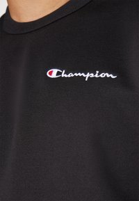 T-shirt en coton noir avec un logo "Champion" brodé en blanc, avec des accents rouge et bleu sur la zone de la poitrine gauche.