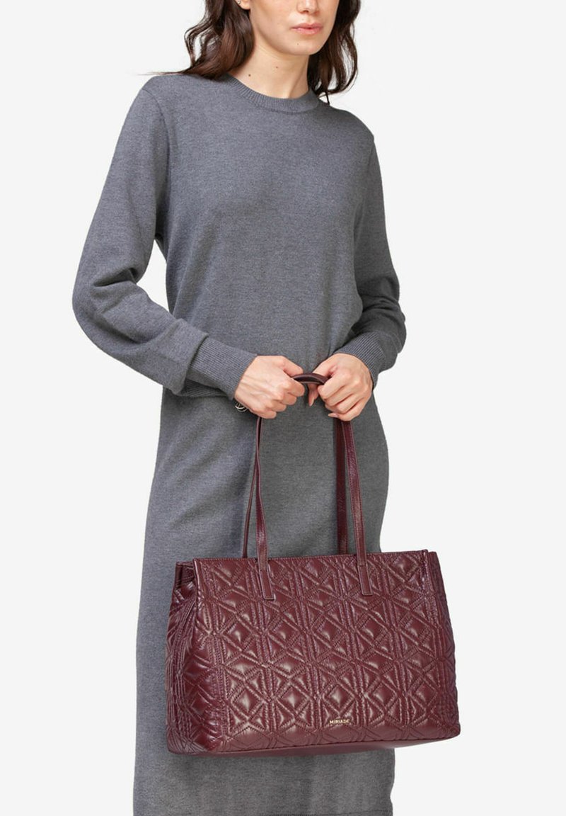 Burgundowa torebka typu tote, pikowana, z wzorem diamentowym, wykonana z błyszczącego materiału, z dwiema długimi rączkami i sztywną formą.