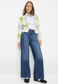 Gebreid vest in crème, groen en blauw met een ruitpatroon. Gecombineerd met hooggerichte, wijd uitlopende spijkerbroek en zwarte sneakers.