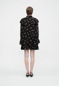 Άτομο που φοράει ένα μαύρο φλοράλ φόρεμα με φρου φρου και μαύρες γόβες, στέκεται με την πλάτη στραμμένη προς μια απλή ανοιχτόχρωμη φόντο.