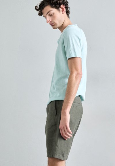 Chemise à manches courtes bleu clair associée à un short vert olive, avec une coupe décontractée et un tissu texturé. Le modèle est représenté de profil.