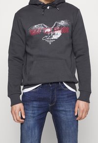 Sudadera gris con un bolsillo delantero que presenta un gráfico de un águila y texto rojo que dice "SOLO LOS VALIENTES". Usada sobre una camiseta blanca y combinada con jeans azules.