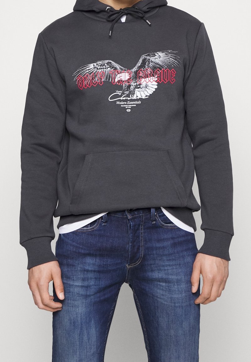 Sudadera gris con un bolsillo delantero que presenta un gráfico de un águila y texto rojo que dice "SOLO LOS VALIENTES". Usada sobre una camiseta blanca y combinada con jeans azules.