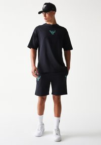 Μαύρο βαμβακερό μπλουζάκι με λογότυπο ταύρου σε τιρκουάζ χρώμα στο στήθος, συνδυασμένο με μαύρο σορτς με αντίστοιχο λογότυπο και λευκά αθλητικά παπούτσια.