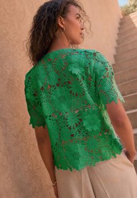 Top verde din dantelă cu modele florale și mâneci scurte, asortat cu pantaloni bej deschis, evidențiat pe un fundal texturat.