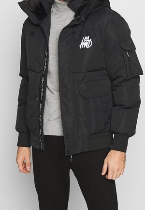 Homme portant une doudoune noire avec un logo blanc sur la poitrine, zippée ouverte sur un col roulé gris clair et un pantalon noir.